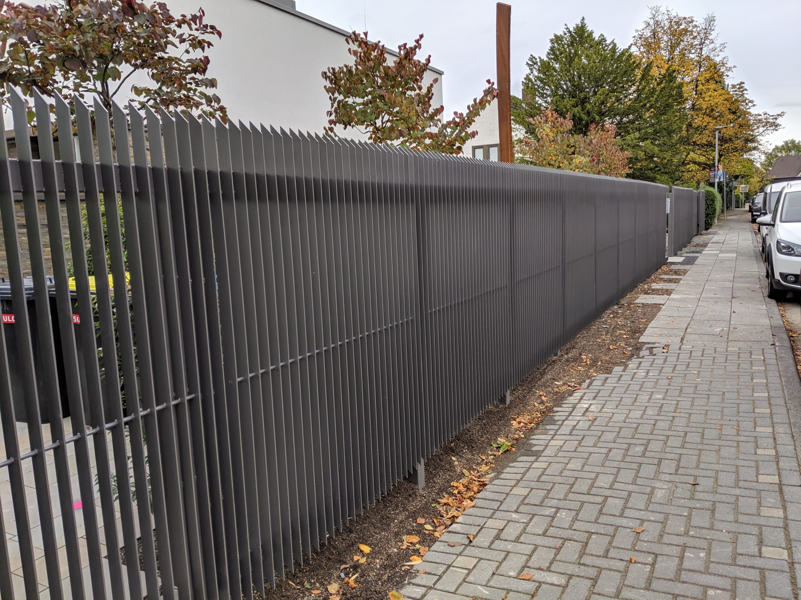 Lange graue Zaunanlage aus vertikalen Stahlprofilen entlang eines Wohnstraßenabschnitts.