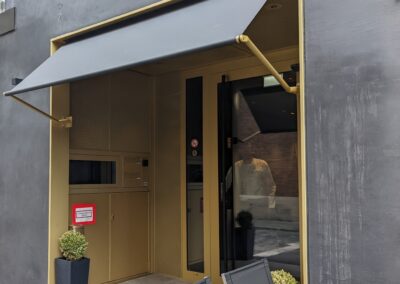 Hoteleingang mit glatten Blechpaneelen, Löschwasseranschlussschrank und strukturierten Streckmetallflächen